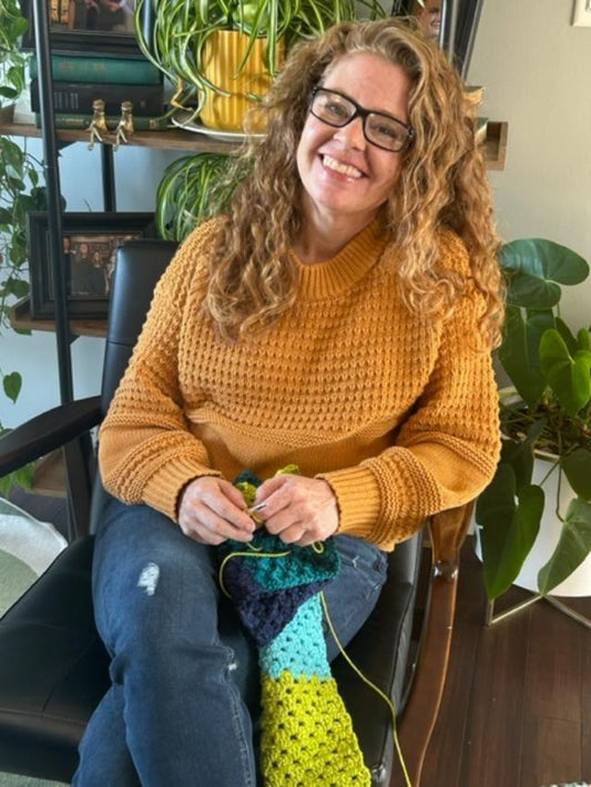 Christy Goble Sits with her crochet needles and plants in the background